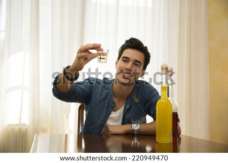 Young man sitting drinking alone at a table, making a toast, with two bottles of liquor alongside him sipping from shot glass