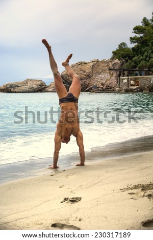 Fit muscular man in his swimsuit doing a handstand on the beach at the edge of the surf as he enjoys a summer day at the coast