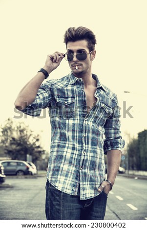 Trendy cool young man standing outside smoking, lowering sunglasses, wearing shirt and jeans