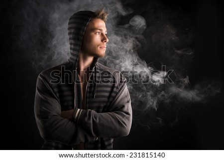 Portrait of handsome tough young man in dark hoodie on black background, looking to a side. Smoke behind him