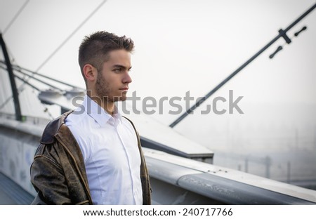 Handsome trendy young man standing on a sidewalk wearing a fashionable jacket and scarf in a relaxed confident pose looking away to a side