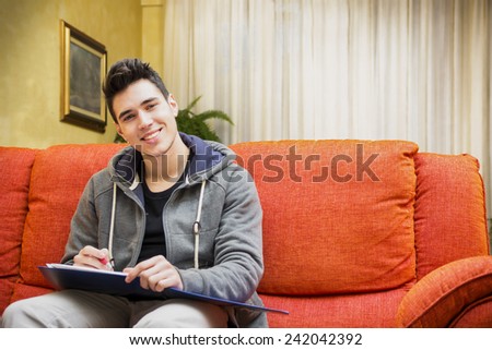Smiling young man at home writing on notebook, sitting on couch, looking at camera