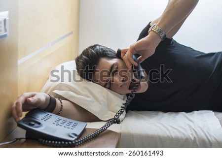 Young Sleepy Man, Lying on his White Bed, Yawning While Talking to Someone Through the Telephone