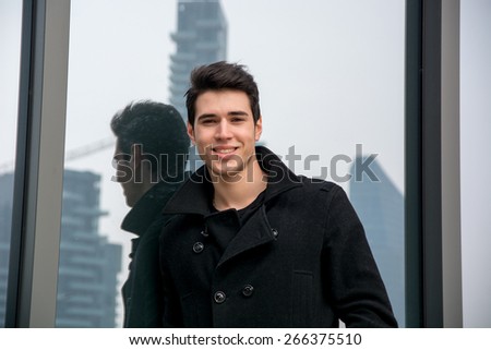Stylish trendy young man standing outdoor against office window, looking confindent at camera