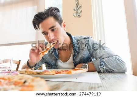 Handsome young man wearing casual denim shirt eating pizza at home, low angle perspective
