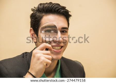 Handsome young man holding a magnifying glass to his eye with a happy friendly smile conceptual of a detective searching for clue