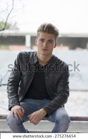 Attractive blond young man in city environment, sitting on handrailing, looking at camera