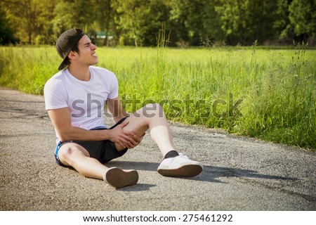 Handsome young man injured while running and jogging on road in the country in a sunny day, wearing white shirt and baseball cap