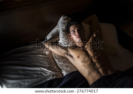 Handsome shirtless athletic young man laying in bed at night looking at camera, seen from above