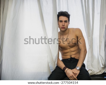 Handsome fit athletic young man shirtless, sitting on window ledge looking at camera