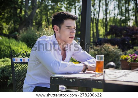 Handsome young man sitting alone at table outside in park or nature, with glass of soda beverage