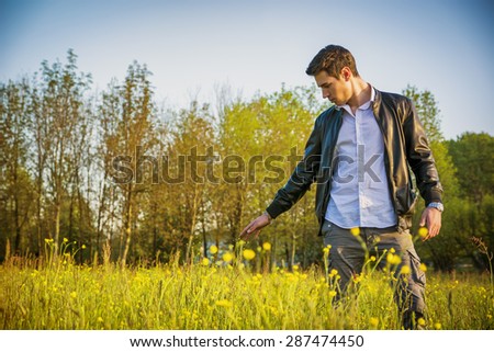 Handsome young man at countryside, touching grass and flowers, standing in  field or grassland, wearing white shirt and jacket, looking down