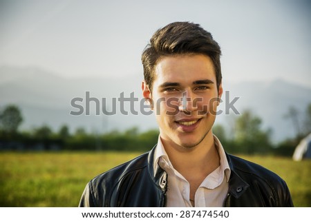 Handsome young man at countryside, in front of field or grassland, wearing white shirt and jacket, smiling and looking at camera