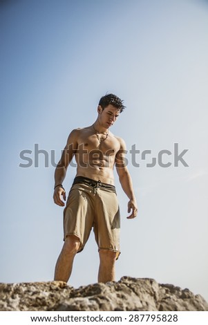 Muscular young man shirtless standing on rock against the sky, seen from below perspective