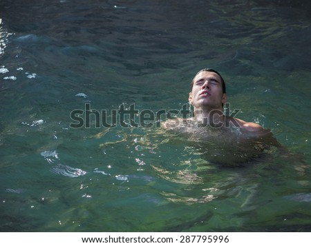 Attractive young shirtless athletic man emerging from water by sea or ocean shore, eyes closed
