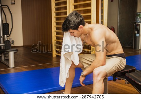 Muscular shirtless male athlete drying sweat from his face with a towel after workout in a gym