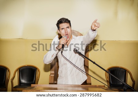 Determined Young Man Speaking Passionately into Microphone and Pointing into Air, Rallying Support for Political Cause in Meeting