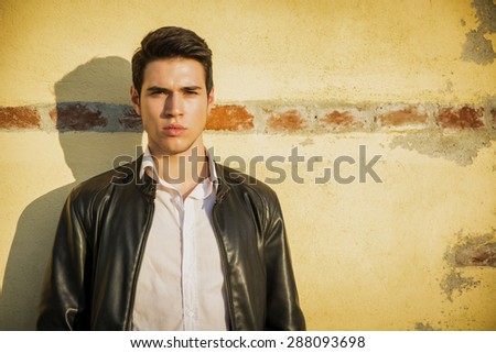 Handsome young man standing in white t-shirt and black leather jacket leaning against old wall behind him, looking at camera