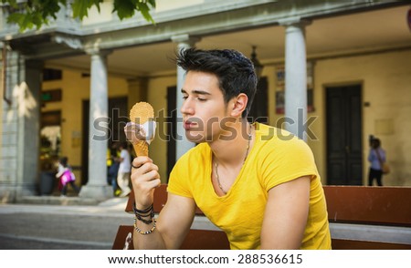Funny attractive young man sitting, wearing yellow T-shirt while eating a tasty vanilla ice cream outdoors in the park, in a warm day of summer