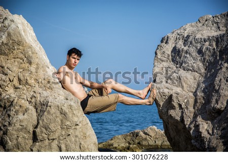 Handsome Shirtless Young Man Wearing Swim Shorts Leaning Across Gap Between Two Massive Boulders at Picturesque Ocean or Sea Coast