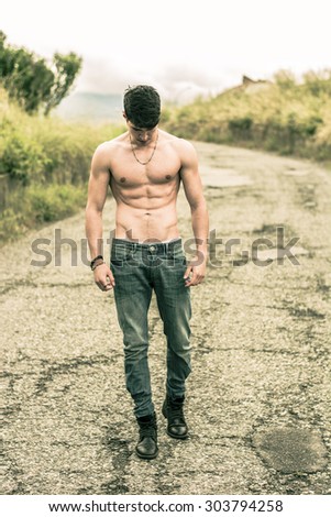 Shirtless sexy muscular young man in jeans walking along rural road, looking down