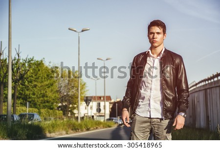 Sexy handsome young man in jeans and black leather jacket walking along city street