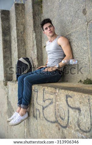 Young Man with Backpack and Bottled Water Lying on Ground Near Staircase - Trick, Impossible Illusion, Image Rotated to Appear as if Man is Sitting on Stone Wall