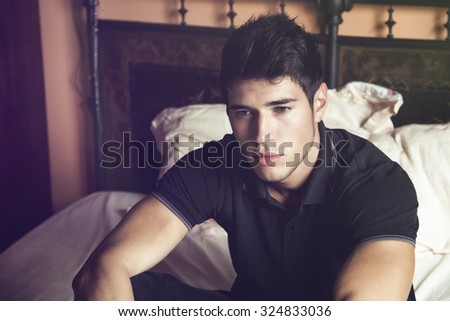 Handsome young man sitting on his bed with serious expression, staring in the void with a blank look