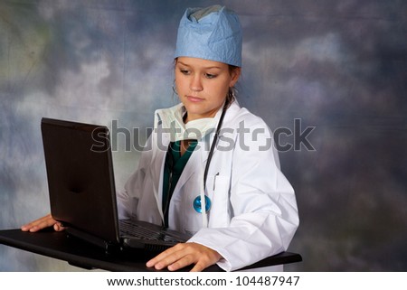 Female medical person in white lab coat, surgery hat and surgery mask around her neck, with a stethoscope around her neck, at a laptop computer