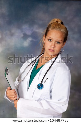 Female medical person in white lab coat, and scrubs, holding a clipboard and looking at the camera with a serious, thoughtful expression