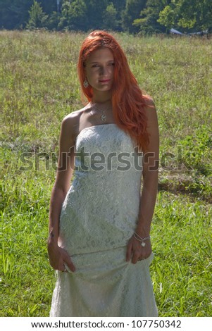 Lovely redhead woman standing outside in a green field, in her white wedding dress