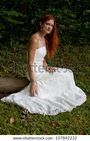 Lovely redhead woman in a white wedding dress, sitting in a field of green grass with woods in the background