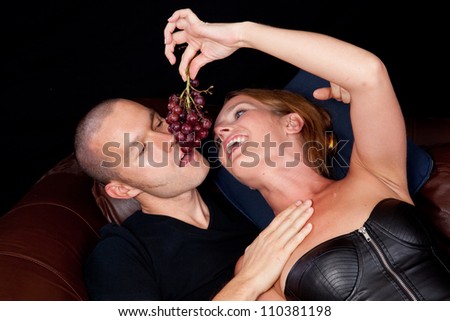 Couple reclining on a couch together, she is feeding him grapes by danling them for him to bite