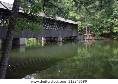 Old wooden covered bridge crossing water with green trees and bushes on the other side.