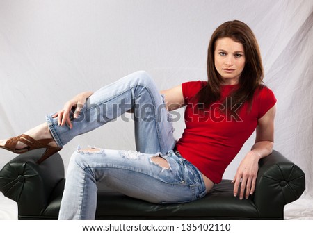 Lovely Caucasian woman in a red shirt and blue jeans, reclining on a bench and looking at the camera with a friendly, content expression