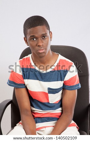 Handsome black man in striped shirt sitting in a chair for a desk, looking at the camera with a thoughtful expression