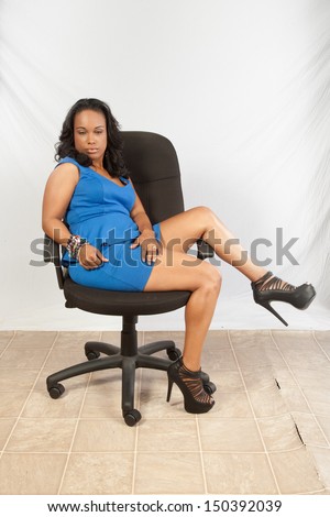 Pretty black woman in blue dress, sitting in a business chair sideways and looking at the camera  with a thoughtful expression