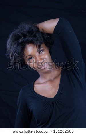 Thoughtful black woman wearing a black shirt shirt,  looking at the camera with one hand behind her head, and a black background