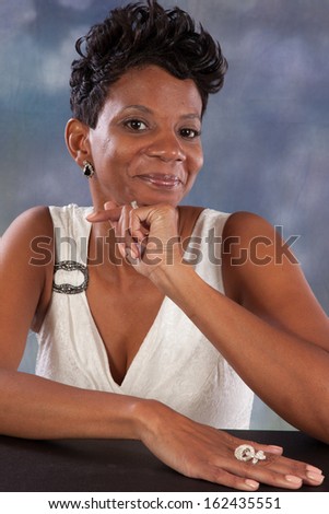 Pretty black woman sitting in a white dress, her chin on her hand and looking at the camera with a friendly, happy smile