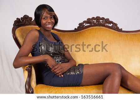 Black woman in a black dress, sitting on a couch and looking at the camera with a happy, friendly smile