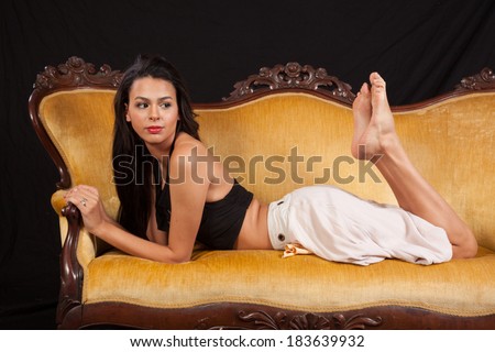 Pretty woman in black top and dress,reclining on a gold couch with her knees bent and feet in the air, and looking at the camera with a serious expression