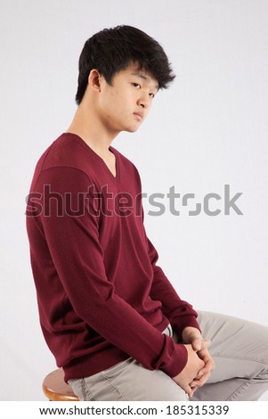 Cute South East Asian teenager  sitting on a wooden stool and looking right with a serious expression.