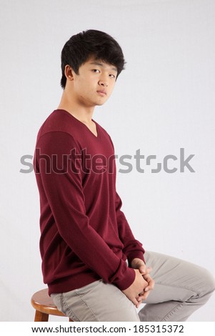 Cute South East Asian teenager in a red sweater,  sitting on a wooden stool and looking  at the camera with a serious expression.