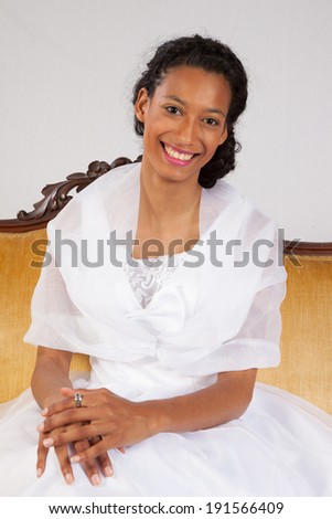 Lovely black woman in white dress, sitting on a gold couch, looking at the camera with a friendly, happy smile