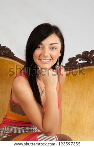 Pretty woman sitting on gold couch with chin on hand looking at the camera with a happy, friendly smile