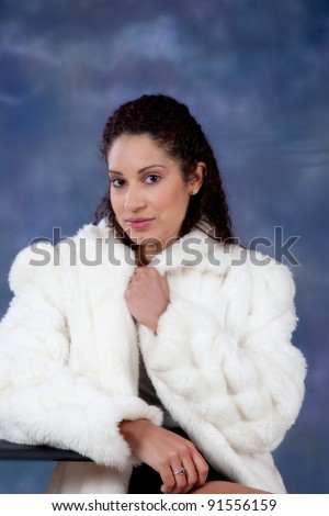 Pretty caucasian woman In a white fur coat looking at the camera and leaning gently on her right arm.