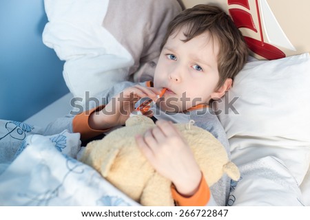 Little boy lying sick in bed. In the mouth, holding a thermometer to measure body temperature. hugs a teddy bear