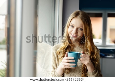 Beautiful smiling blond woman drinking coffee by the window. She looks at the camera