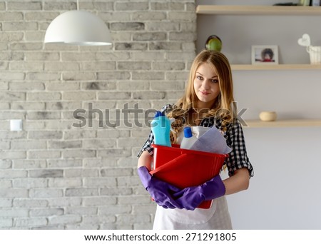 Smiling  blond woman holding a bucket full of cleaners. She is wearing a white apron. On hands has protective rubber gloves