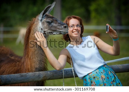Funny selfie with my friend. Attractive smiling young woman holding smartphone and making selfie with lama outdoors